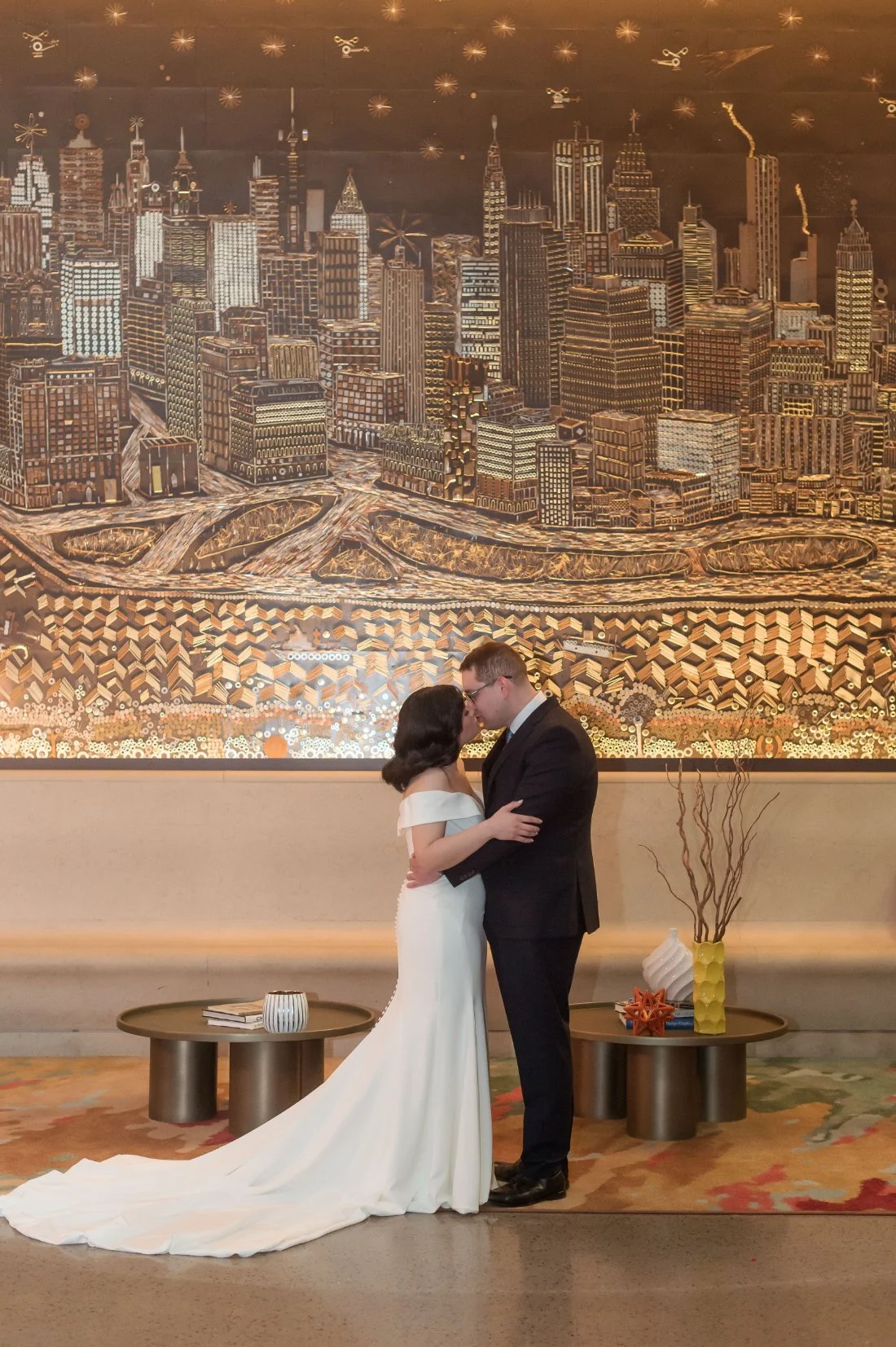   Bride and groom share a quiet kiss in front of a large golden cityscape mural. The bride wears an off-the-shoulder gown with a long train, and the groom wears a dark suit and glasses.   micro wedding photo timeline 