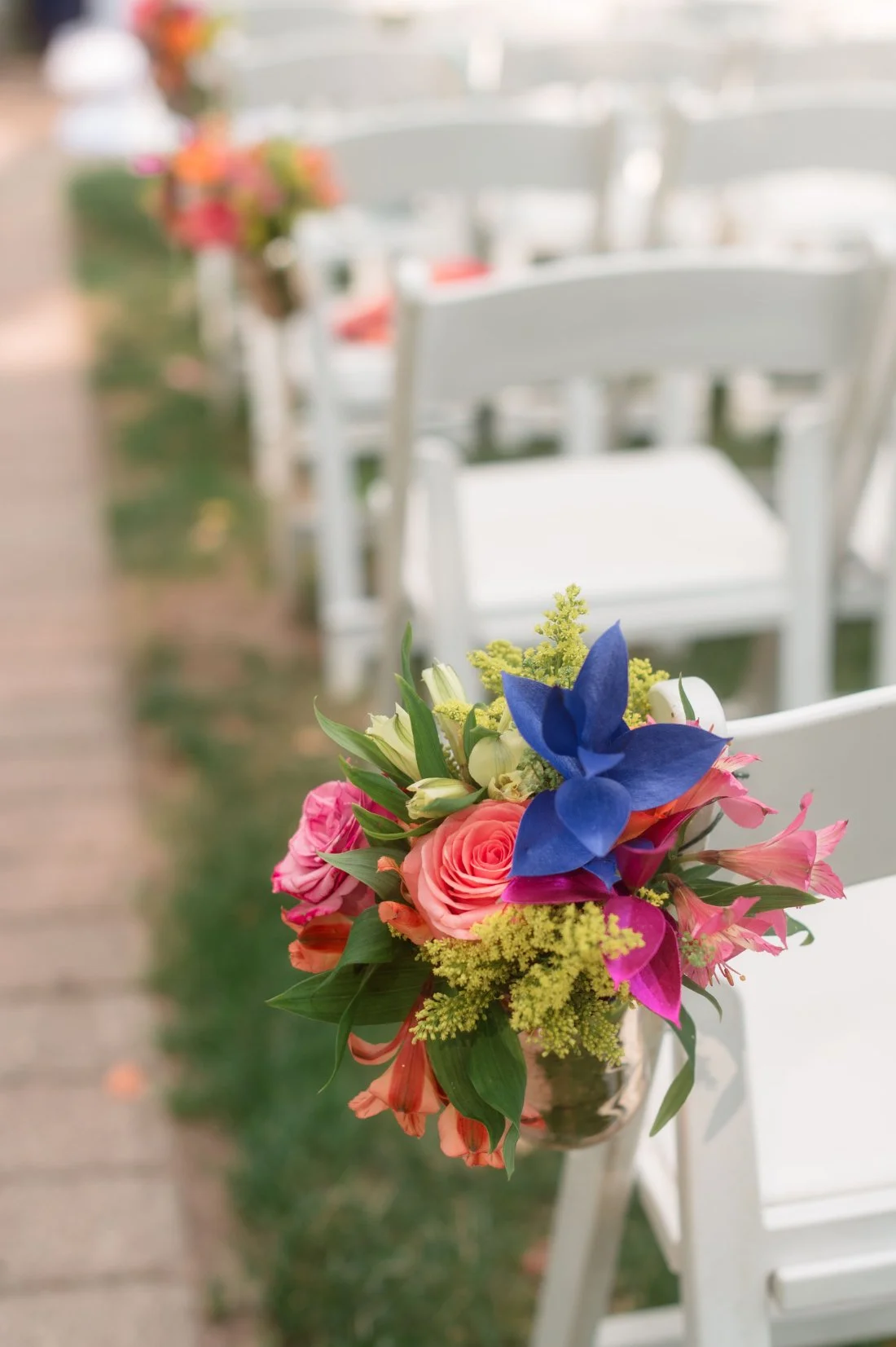   Close-up of a white ceremony chair decorated with a bright floral arrangement, including coral roses, blue orchids, yellow filler, and lush greenery, lining a garden wedding aisle.  