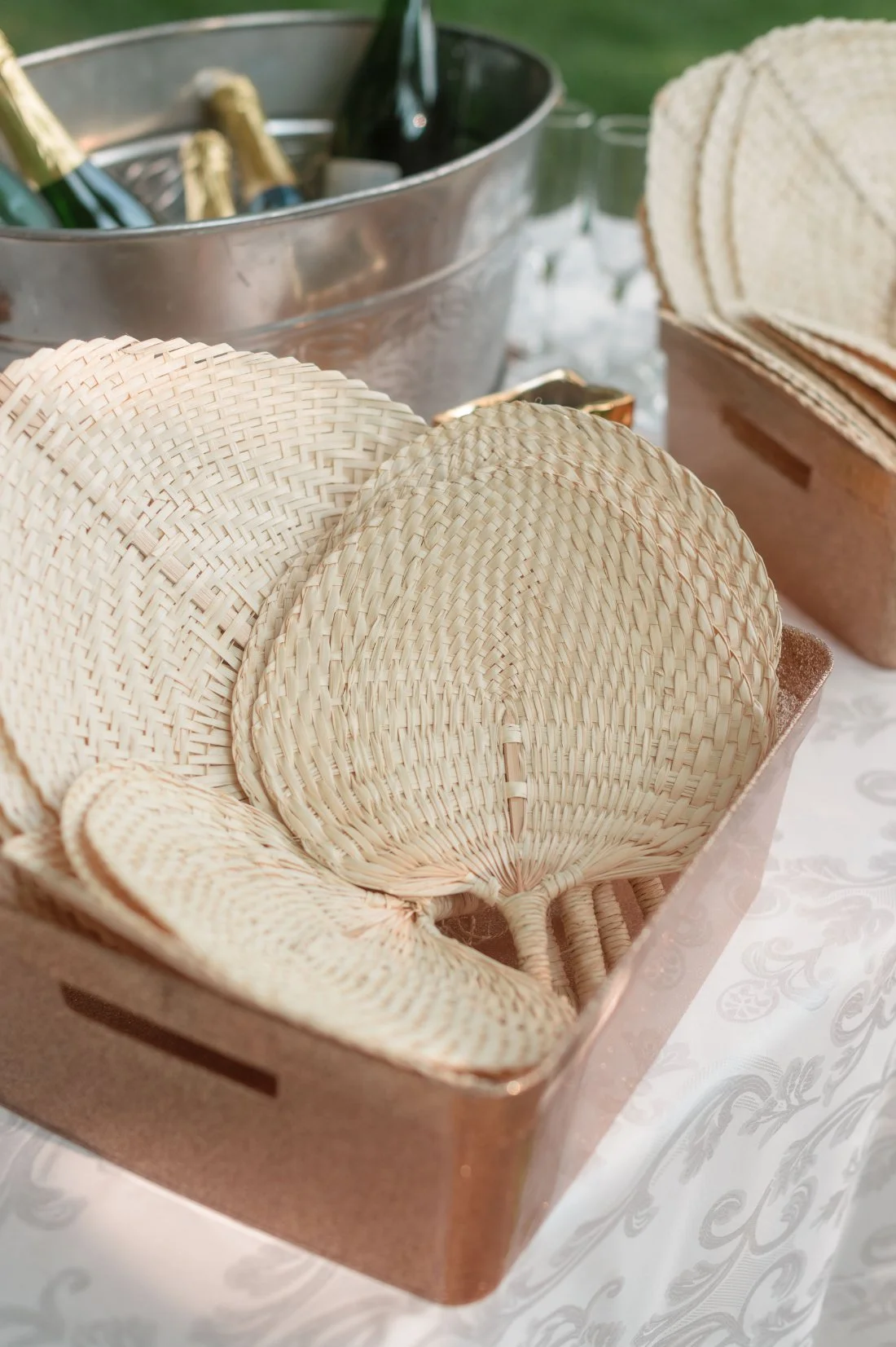   Woven hand fans in a copper-toned basket on a white patterned tablecloth, set next to a large metal tub filled with champagne bottles — a thoughtful outdoor wedding detail for guests.    