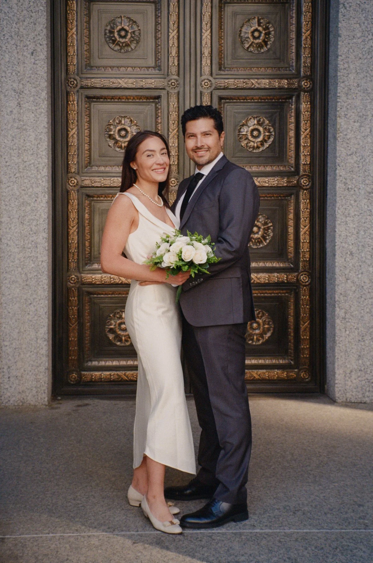  A bride in a simple white dress and a groom in a dark suit smile and embrace in front of ornate bronze doors, holding a small bouquet of white flowers. | wedding photo timeline 