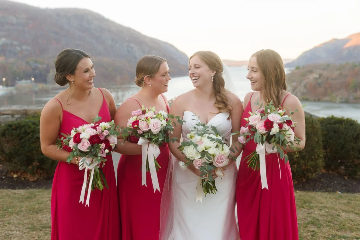  A bride and three bridesmaids in pink dresses stand outdoors by the water, smiling at each other and holding lush pink-and-white bouquets. 