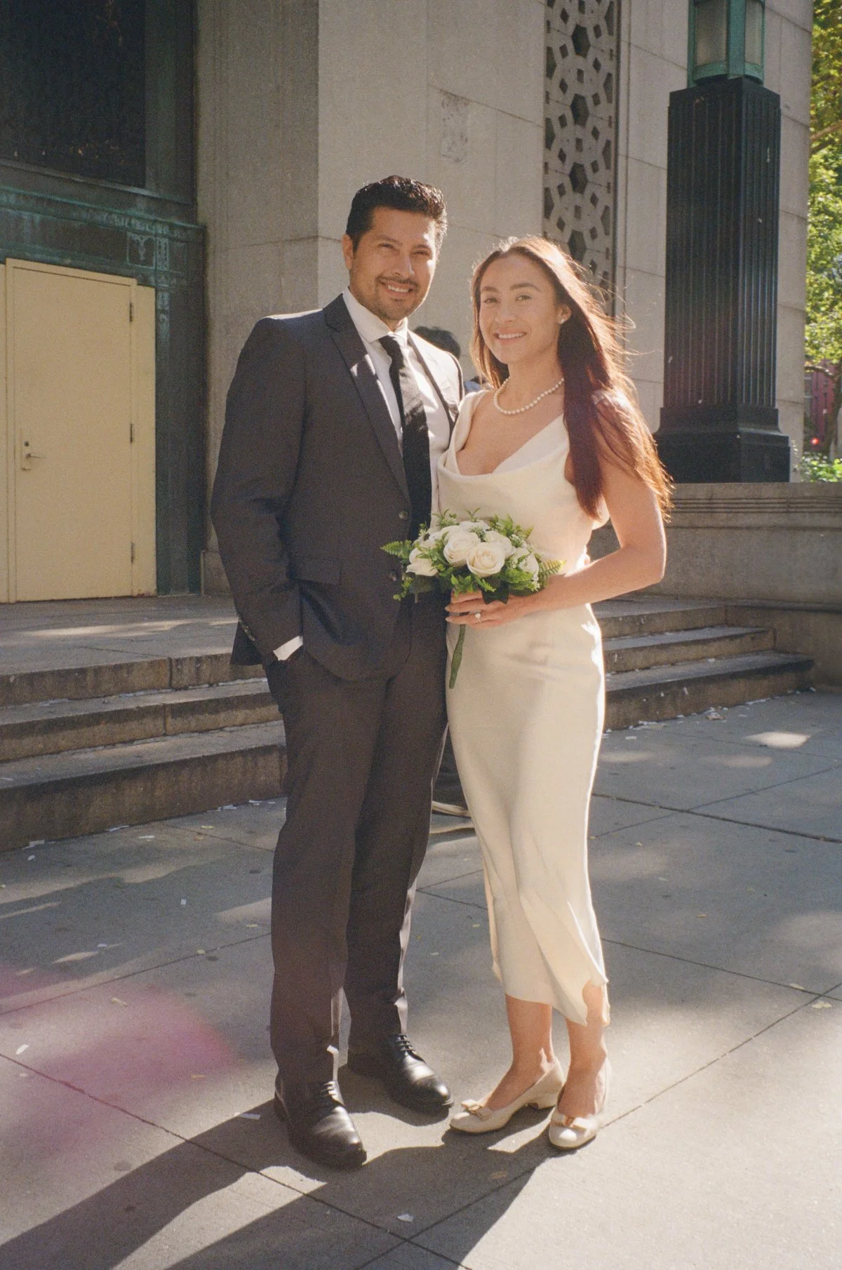 Bride and groom stand outside a grand stone building, smiling in soft sunlight, bouquet in hand, captured in elegant film wedding photography style.