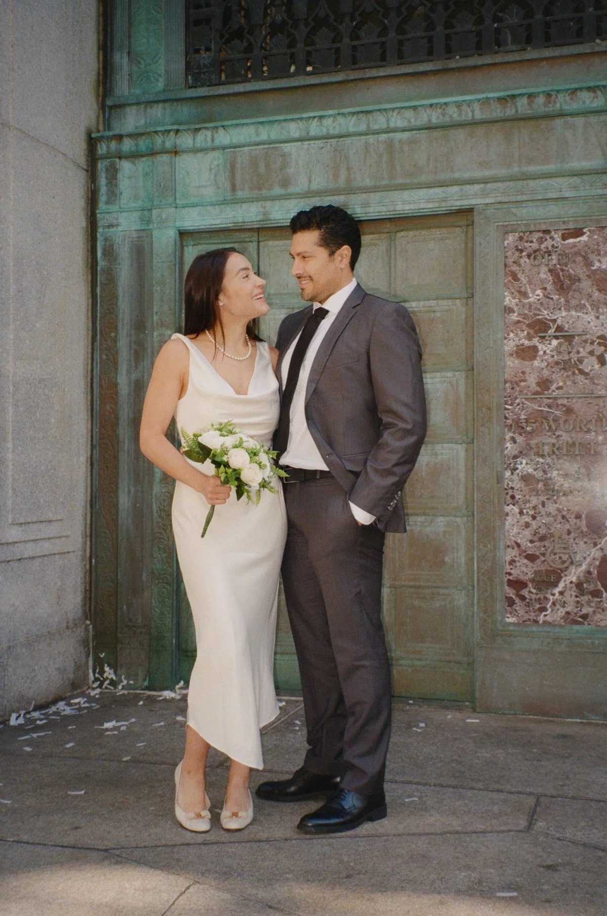  Bride and groom lean close, smiling at each other against an ornate doorway, intimate urban portrait with timeless film wedding photography tones.   