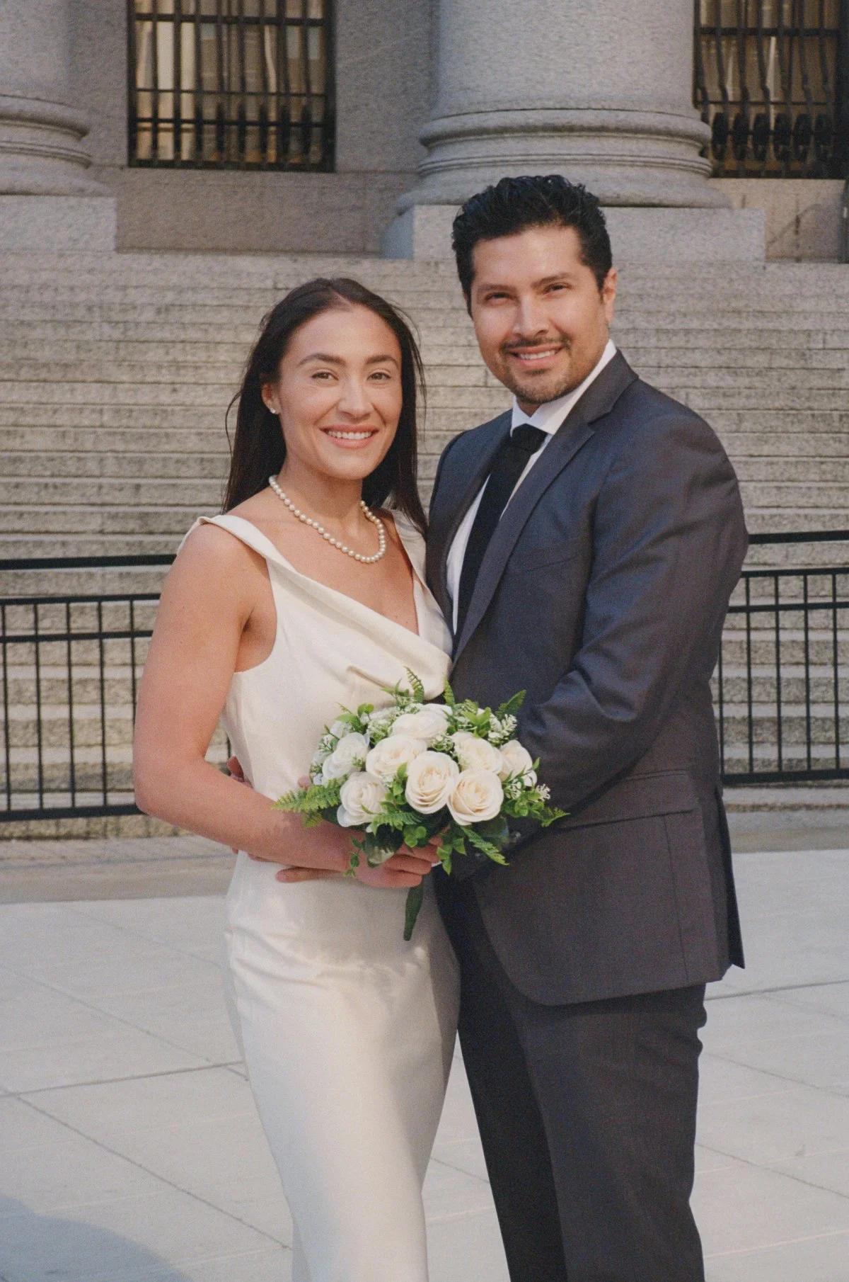  Couple poses together in front of courthouse steps, holding a bouquet and smiling directly at the camera, clean composition in refined film wedding photography.   