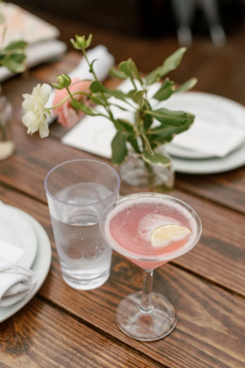  Signature pink cocktail and table florals styled on a wooden reception table during an intimate rooftop wedding celebration. 