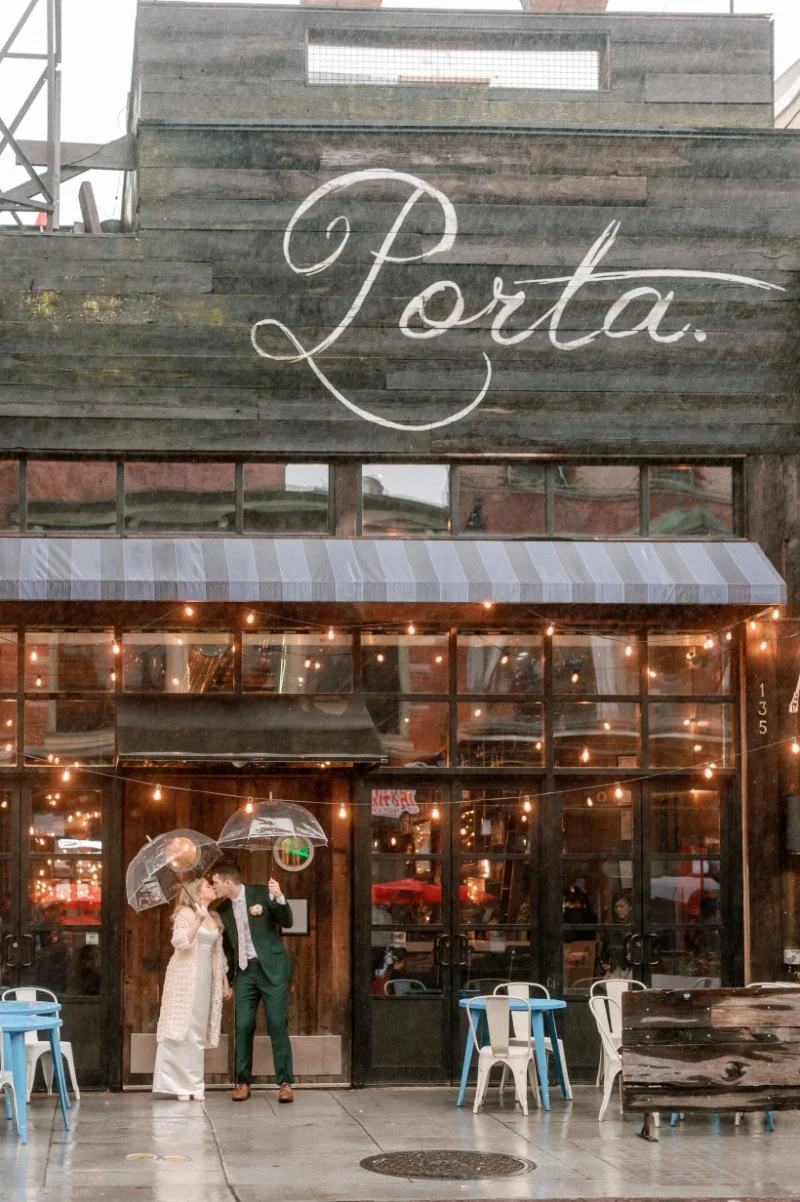  Couple stands outside Porta restaurant in the rain under clear umbrellas after their rooftop wedding in Jersey City. 