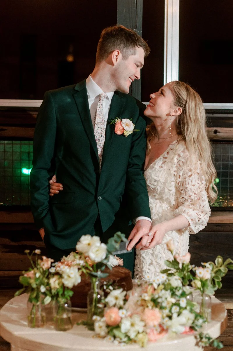  Newlywed couple smiles beside their floral-covered cake table during an intimate rooftop wedding reception in Jersey City. 