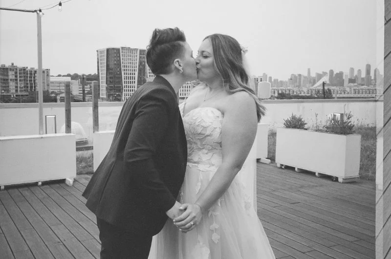  Couple shares a kiss during their rooftop wedding in Jersey City with skyline views and modern city buildings in the background. 