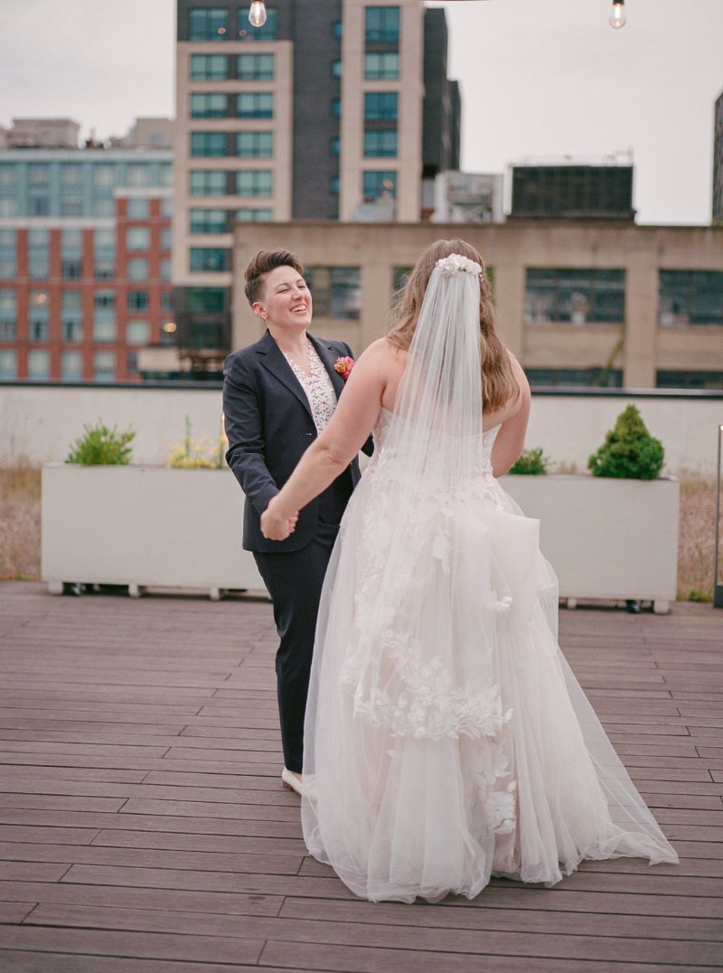  Newly married couple holds hands and laughs together during a rooftop wedding in Jersey City with soft city views behind them. 