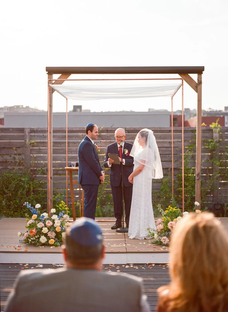  Outdoor wedding ceremony beneath a modern wooden arch with florals during a rooftop wedding in Jersey City at sunset. 