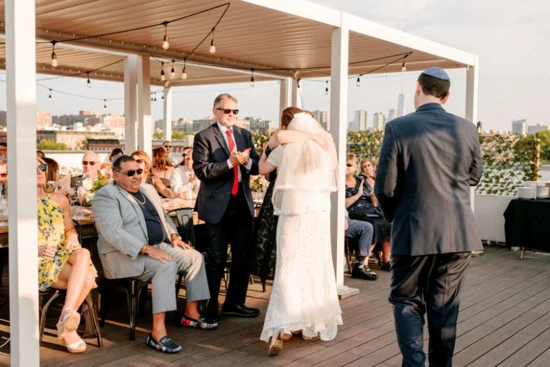  Bride walks down the aisle toward the ceremony space while guests gather beneath string lights on a rooftop venue. 