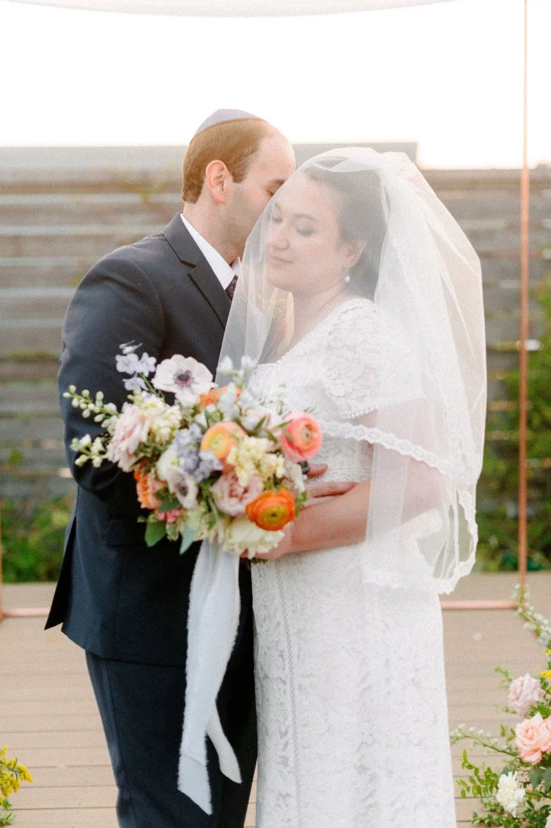 Newlywed couple embraces beneath the bride’s veil while holding a colorful bouquet during their rooftop wedding ceremony. 