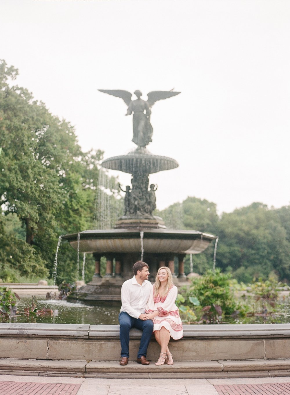  Couple sitting by Bethesda Fountain in soft romantic outfits for timeless engagement photo outfit inspiration. 