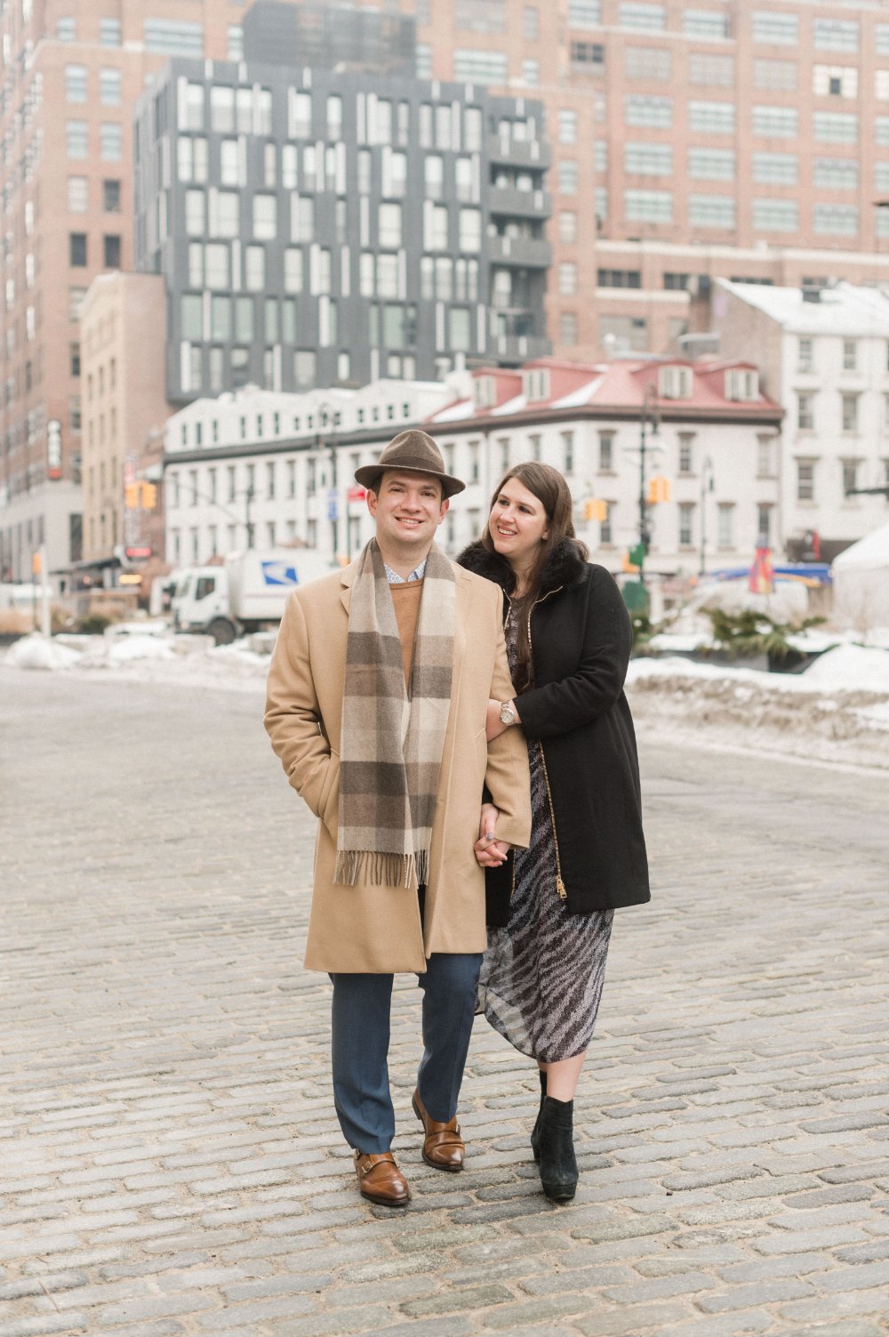  Couple in stylish winter coats walking through the city during a cozy urban engagement session. 