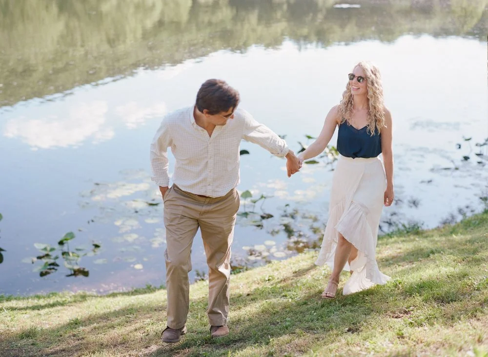  Couple walking hand in hand beside the lake during a relaxed summer engagement session. 