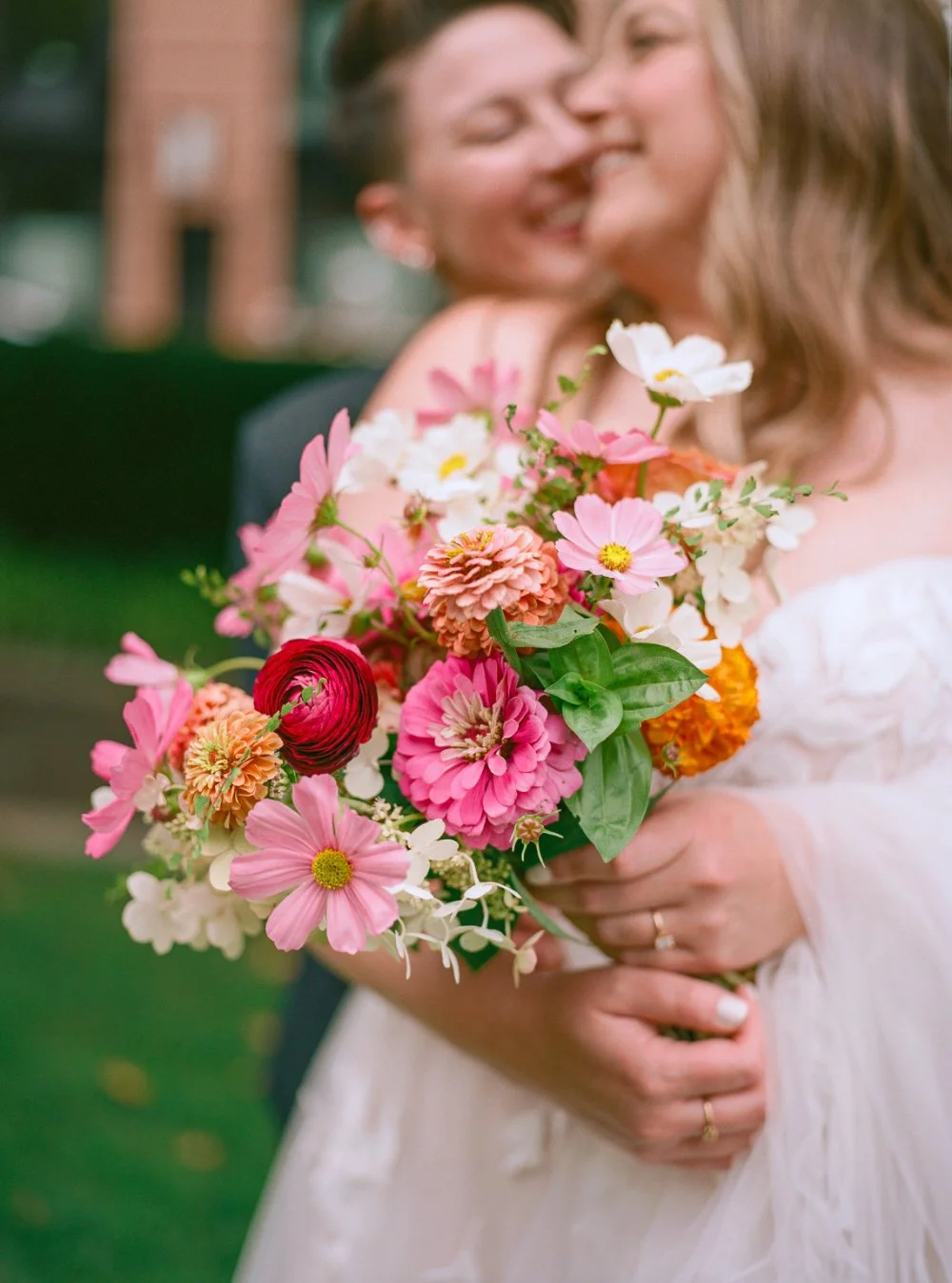 LGBTQ wedding photographer captures colorful bridal bouquet with couple embracing softly in the background. 
