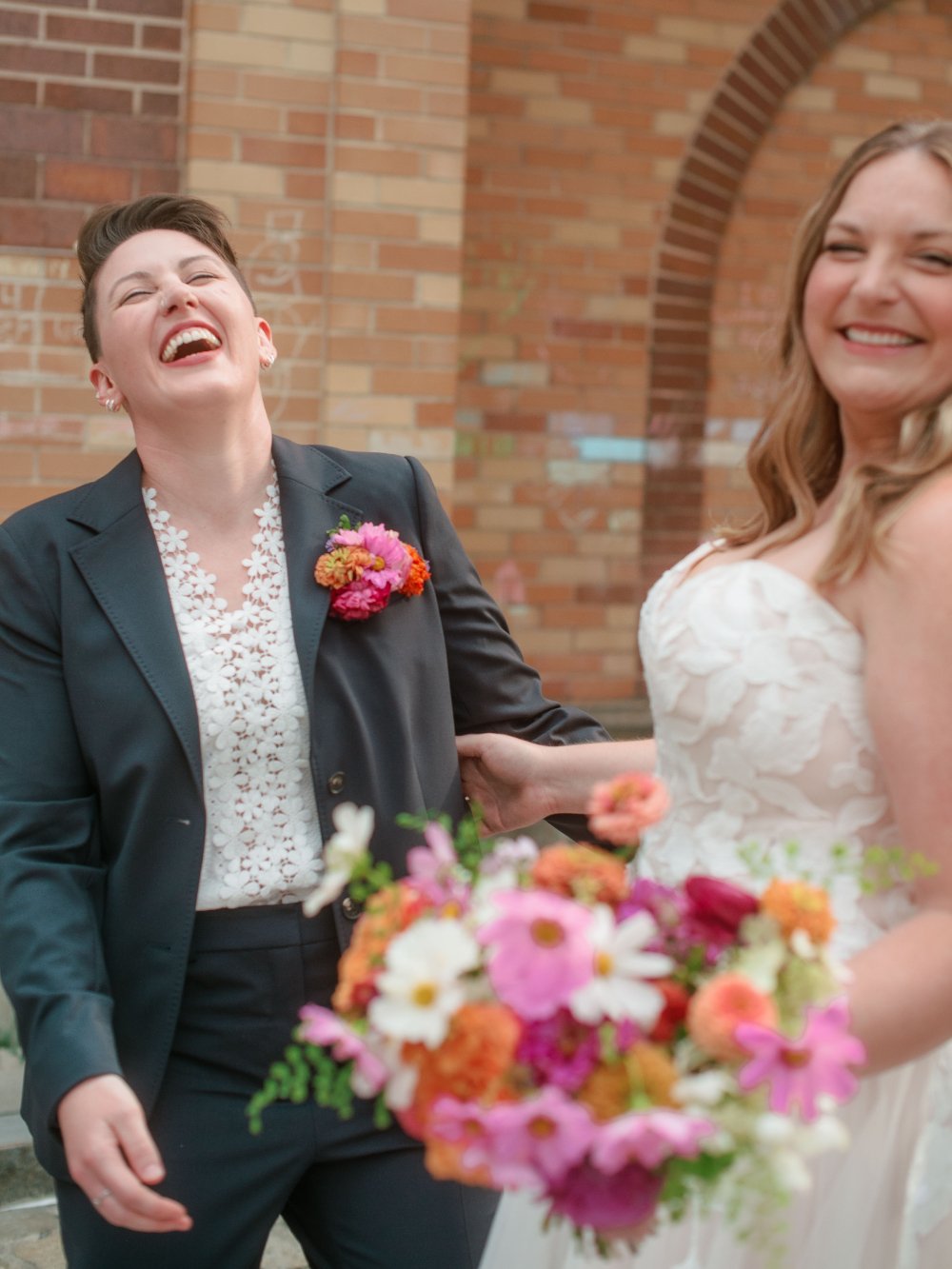  LGBTQ wedding photographer captures joyful newlyweds laughing together while holding colorful wedding bouquets. 