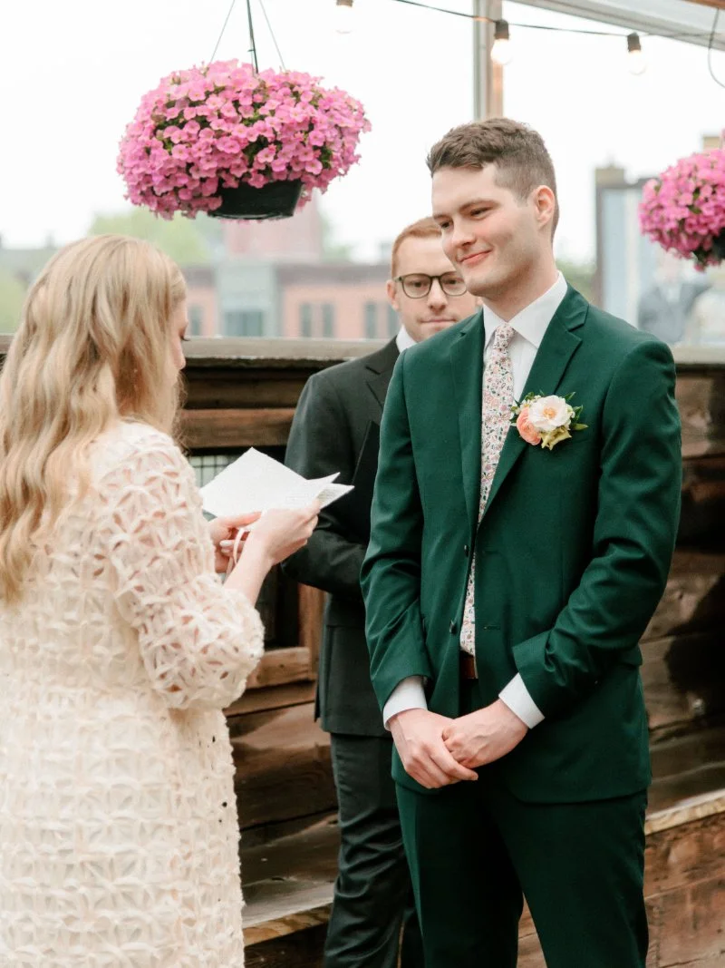  Bride reading her wedding vows as the groom smiles during their intimate rooftop wedding ceremony. 