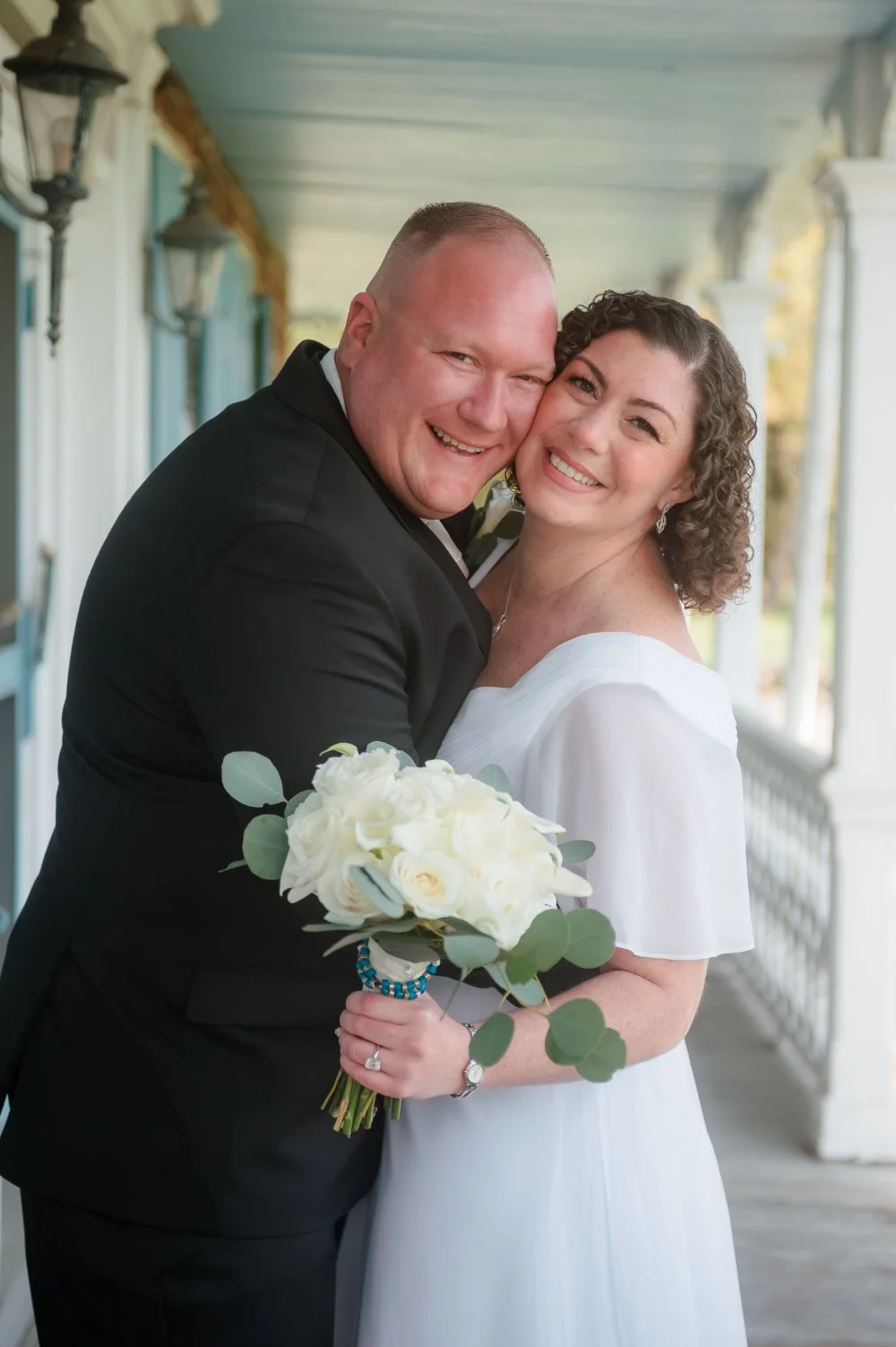  Newlyweds smiling and embracing while holding a white rose bouquet after their wedding ceremony. 