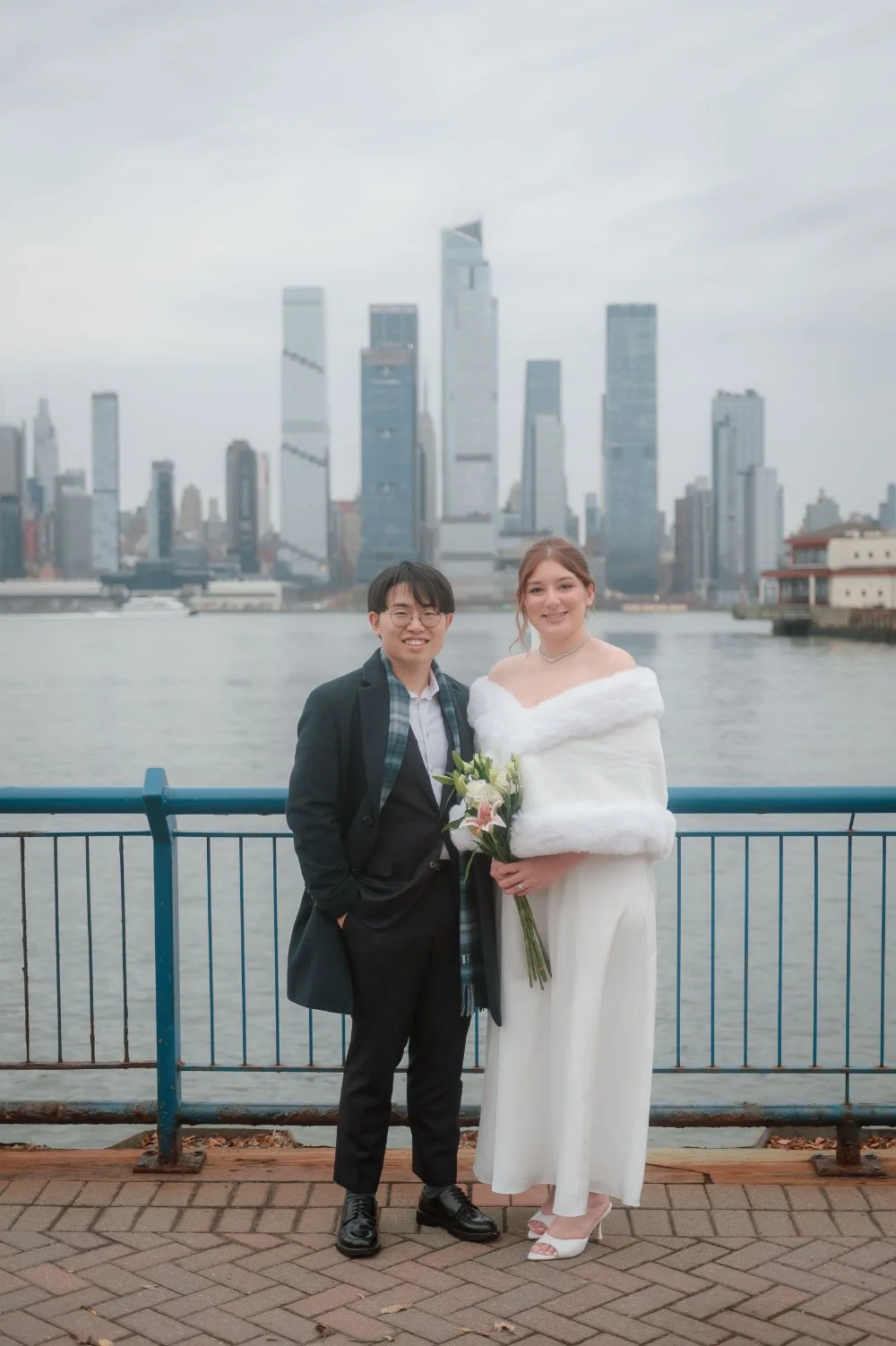 Happy couple posing by the waterfront during their NYC holiday wedding, with the Manhattan skyline behind them and the bride holding a bouquet of lilies, offering romantic winter wedding inspiration for New Jersey and NYC couples. 