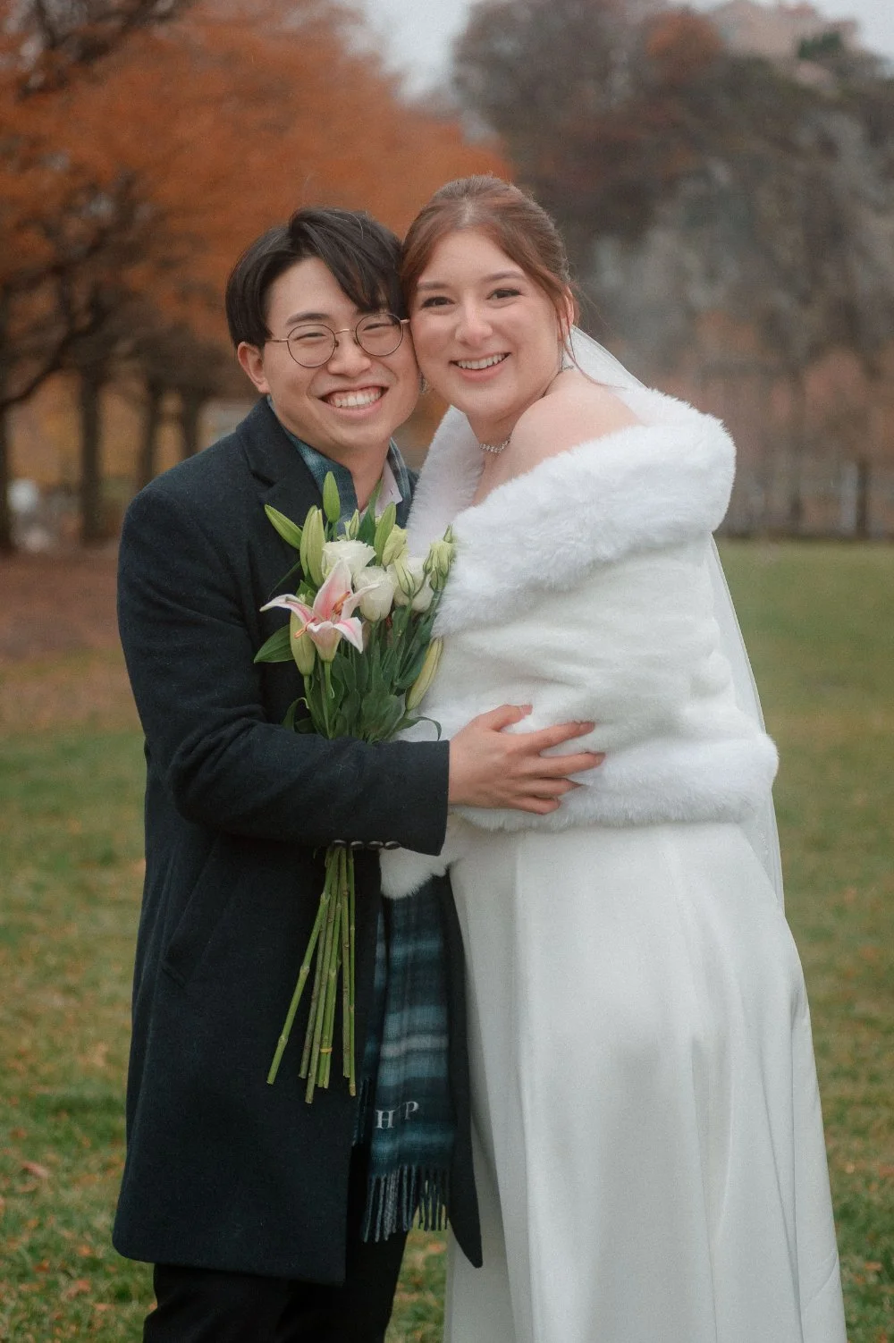  Smiling newlyweds embracing outdoors during their NYC holiday wedding, with the bride wearing a white fur wrap and holding a bouquet of pink and white lilies, offering romantic winter wedding inspiration for New Jersey and NYC couples. 
