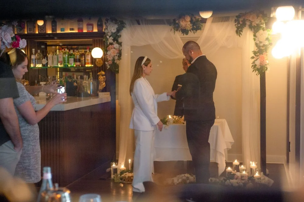  Newlyweds exchanging rings during their intimate holiday wedding, surrounded by white flowers, greenery, and candlelight in a cozy setting inspired by NYC and New Jersey weddings. 