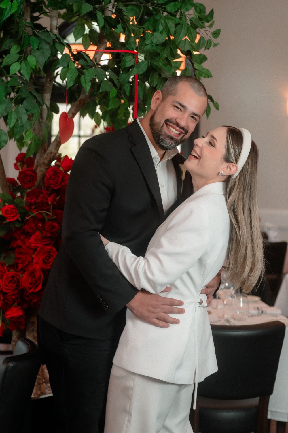  Happy couple embracing during their intimate holiday wedding, surrounded by red roses, lush greenery, and warm lighting in NYC, New Jersey wedding inspiration. 