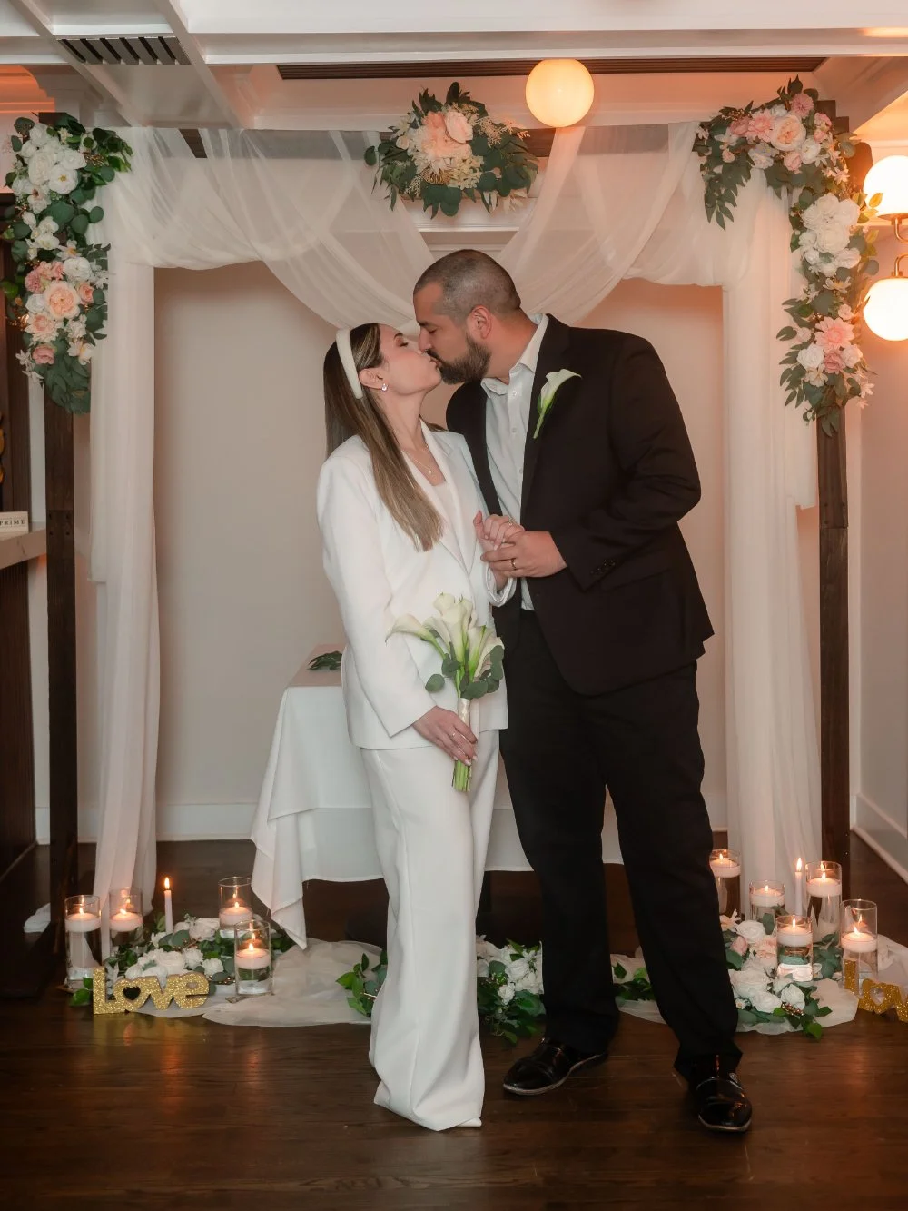  Newlyweds sharing a romantic kiss during their intimate holiday wedding, surrounded by white flowers, greenery, and candlelight in a cozy setting inspired by NYC and New Jersey weddings. 