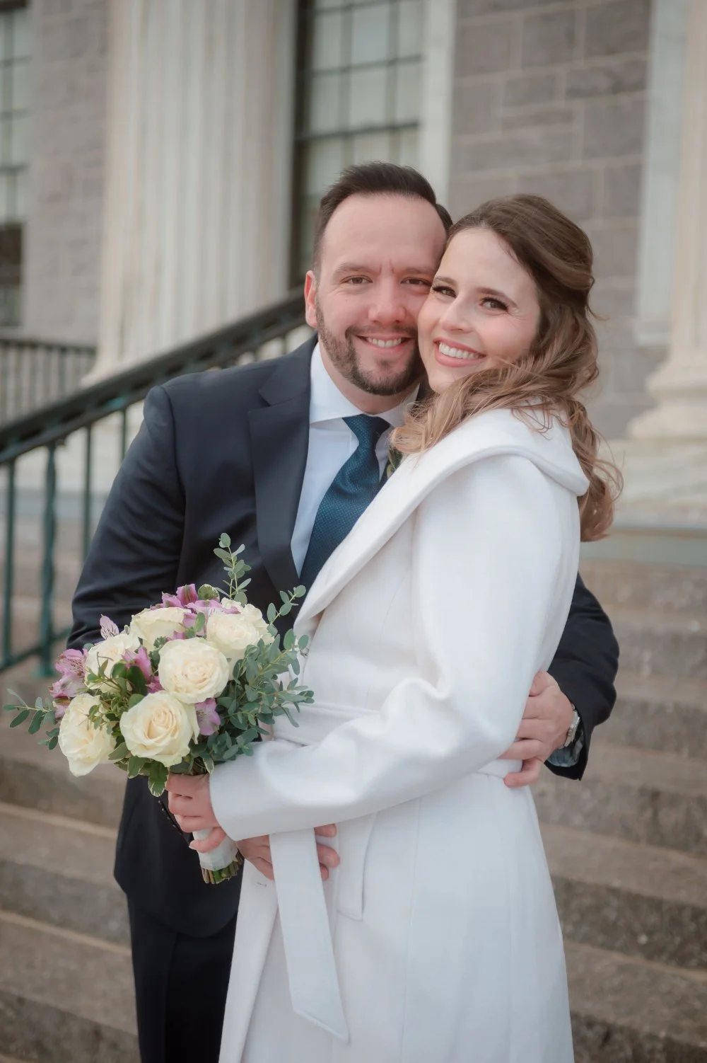  Happy couple embracing outdoors during their intimate holiday wedding, with the bride holding a bouquet of soft pink and white flowers, perfect for NYC and New Jersey wedding inspiration. 