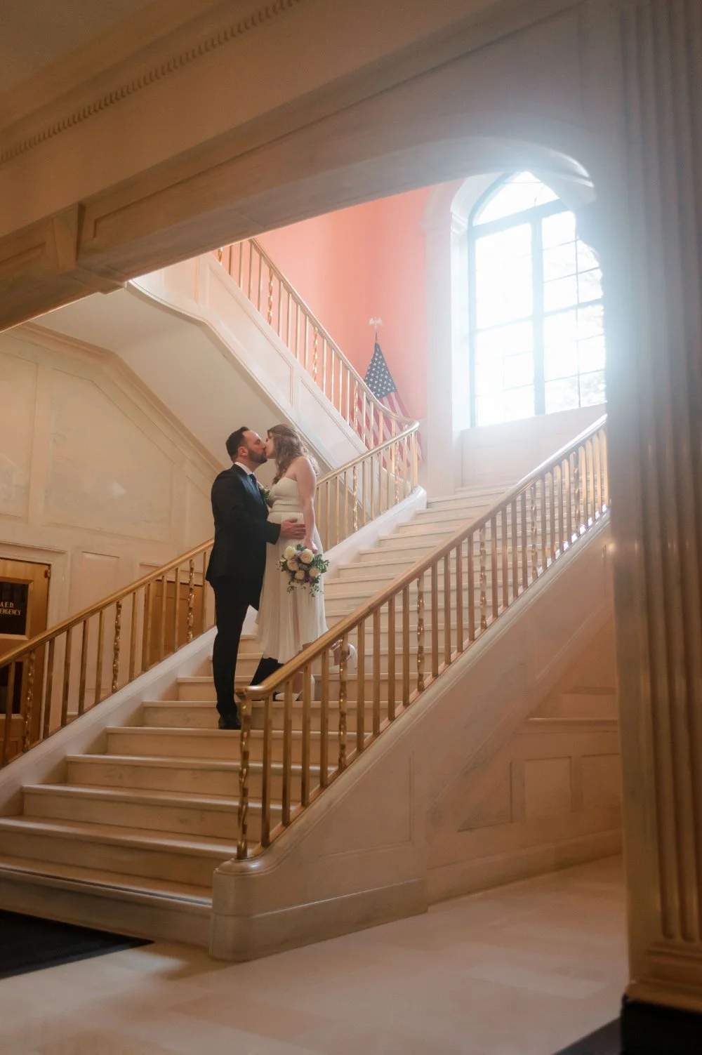  Newlyweds sharing a romantic kiss on a grand indoor staircase during their holiday wedding, surrounded by elegant architecture and natural light, perfect for an intimate NYC or New Jersey celebration. 