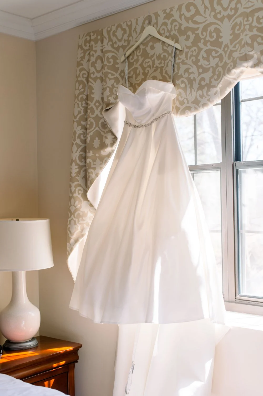 Strapless white wedding dress with a beaded belt hanging beside a bright window.