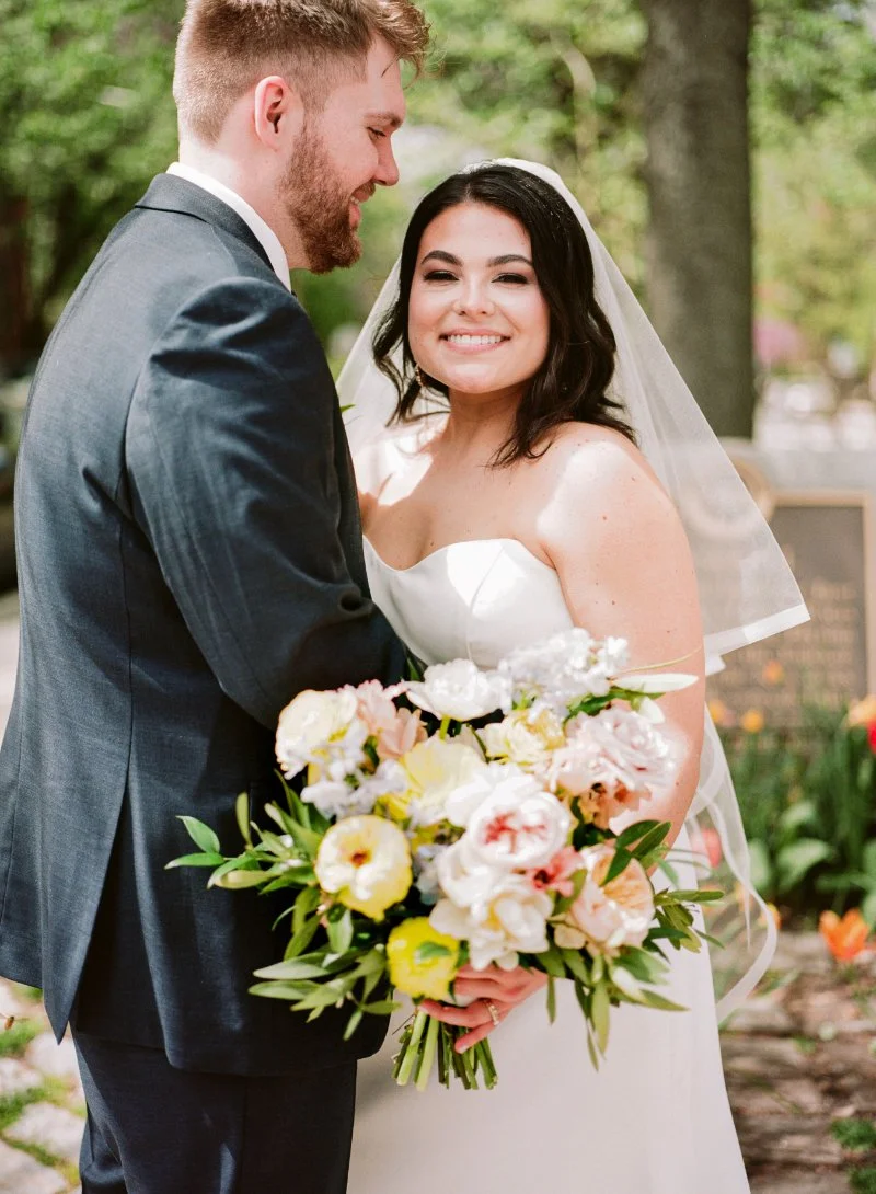 Bride smiling at camera while holding bouquet during outdoor ceremony, supported by a wedding planner for a small wedding.