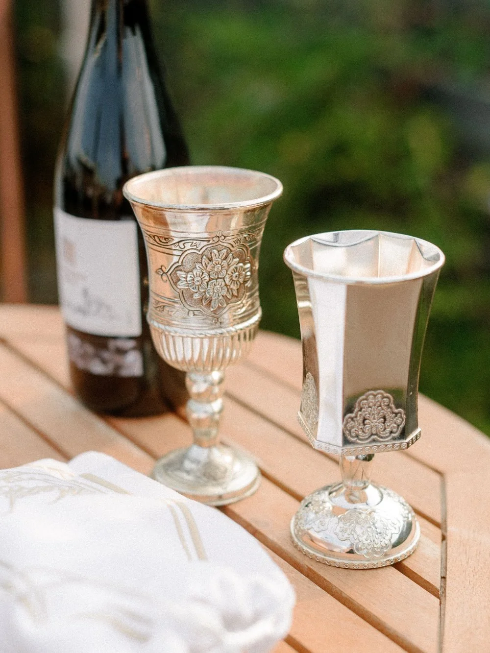 Ornate silver wedding cups displayed beside a wine bottle on a wooden table outdoors.
