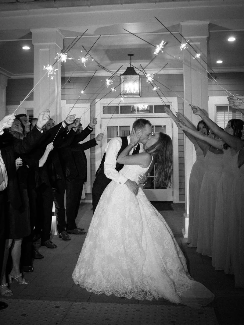 Bride and groom kiss under sparklers during a romantic big wedding exit with their bridal party.