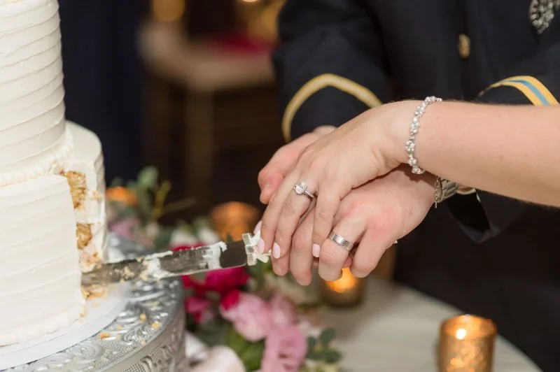 Couple cutting wedding cake together, hands gently placed with rings visible and warm candlelight nearby
