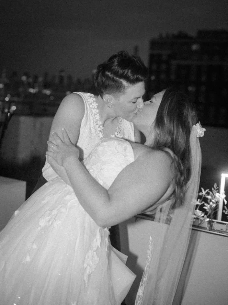 Two brides sharing a kiss during intimate reception, captured in black and white with support from a wedding planner for a small wedding.