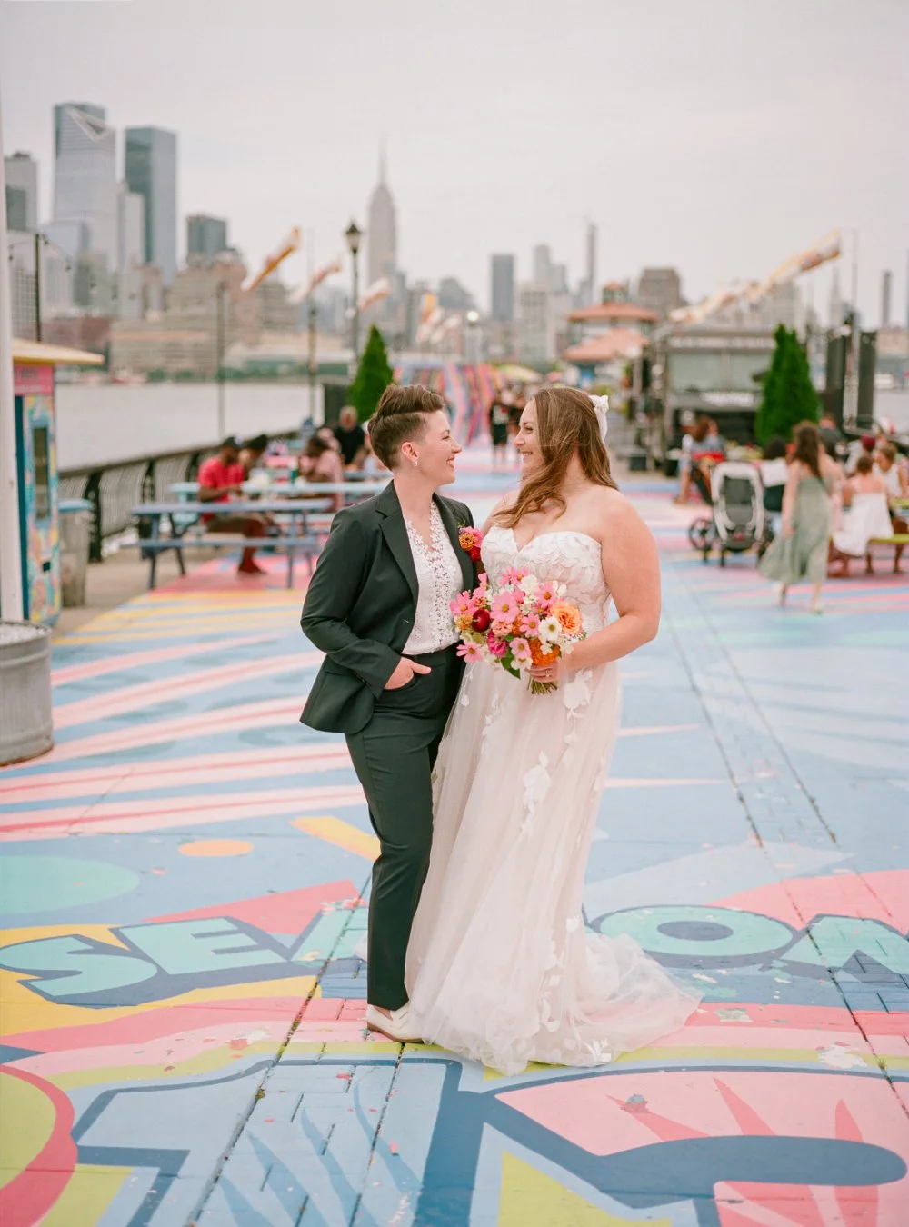 LGBTQ wedding photographer captures newlyweds walking along a colorful waterfront promenade with the New York City skyline behind them.