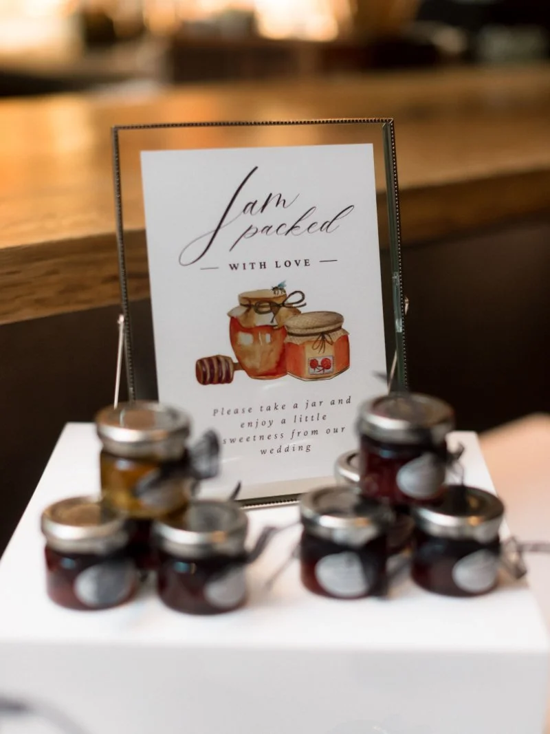 Mini jam jars as favors sit by a sign—sweet keepsakes for guests after the big wedding exit.