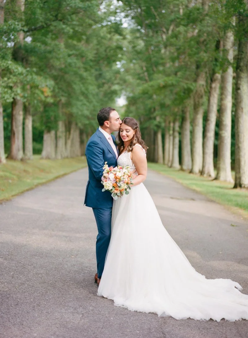 Bride and groom standing on tree lined road while groom kisses bride during romantic outdoor wedding portrait.