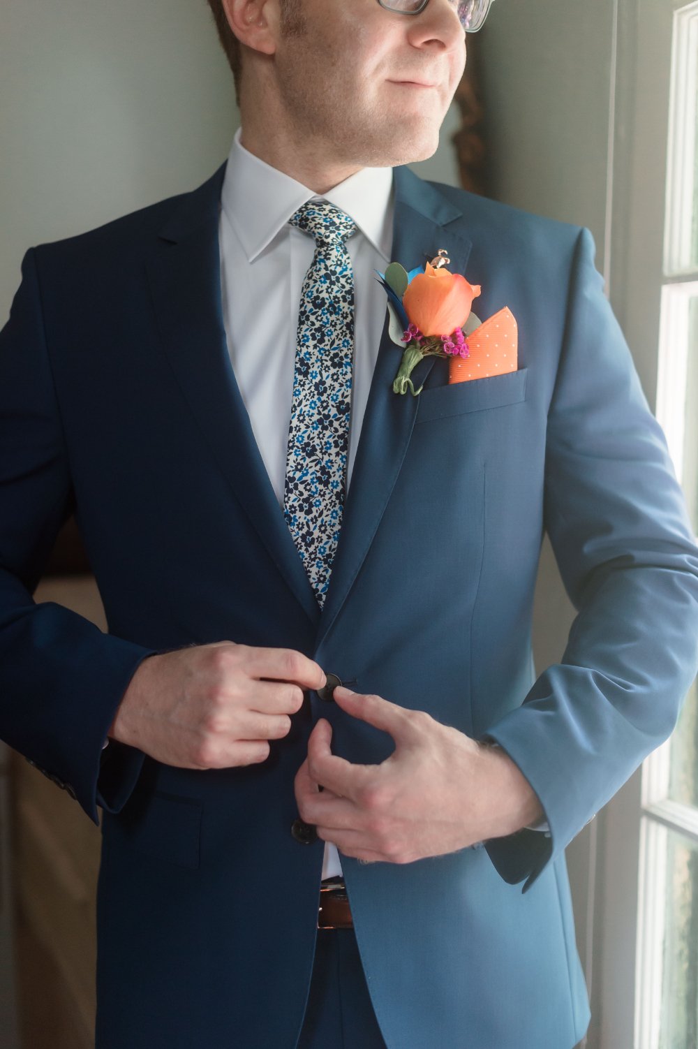 Groom style ideas featuring a navy suit, floral tie, orange pocket square, and colorful rose boutonniere.