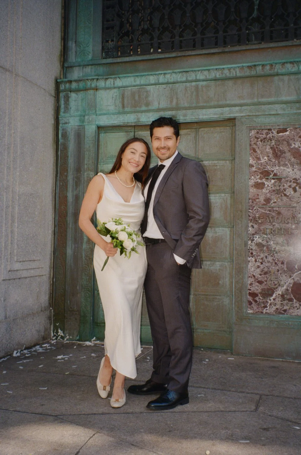 Couple smiling together during their NYC elopement, with the bride wearing a simple white satin dress and the groom in a dark suit, posing outside a historic building.