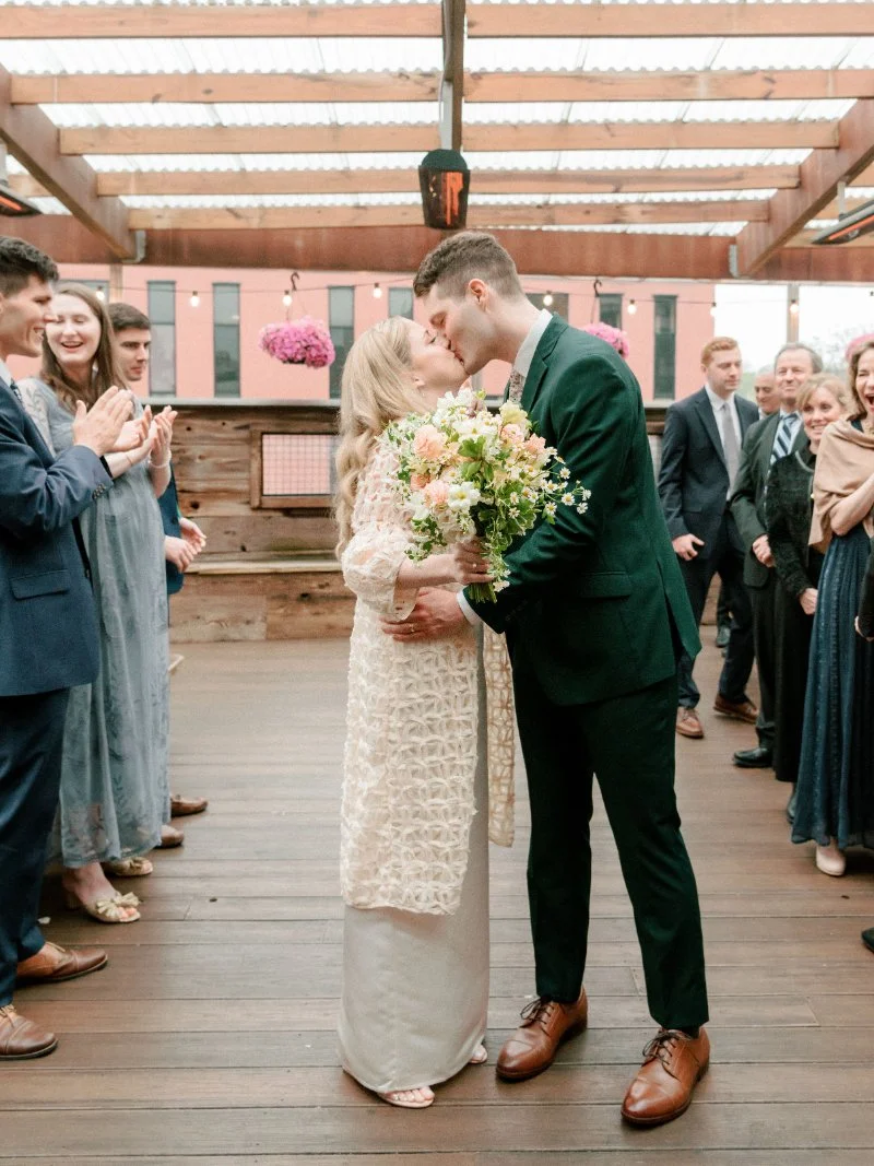 Rooftop wedding ceremony with couple kissing, guests gathered around, soft florals and warm light