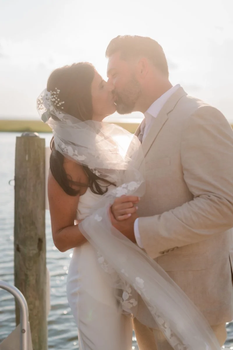 Bride and groom sharing a sunset kiss during golden hour photography beside the water on their wedding day.