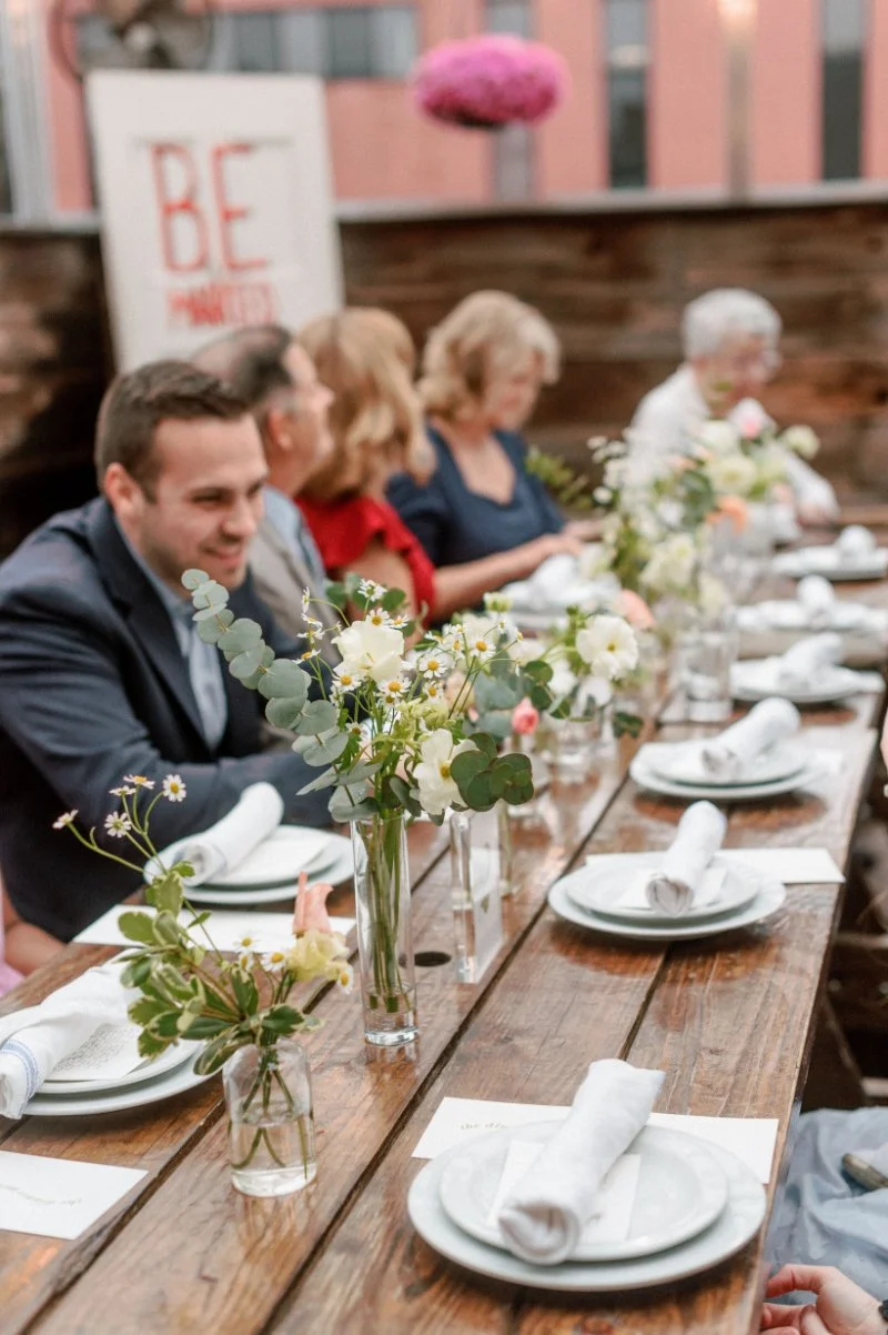 Long wooden table set with simple plates and fresh flowers, guests gathered for a warm, intimate meal