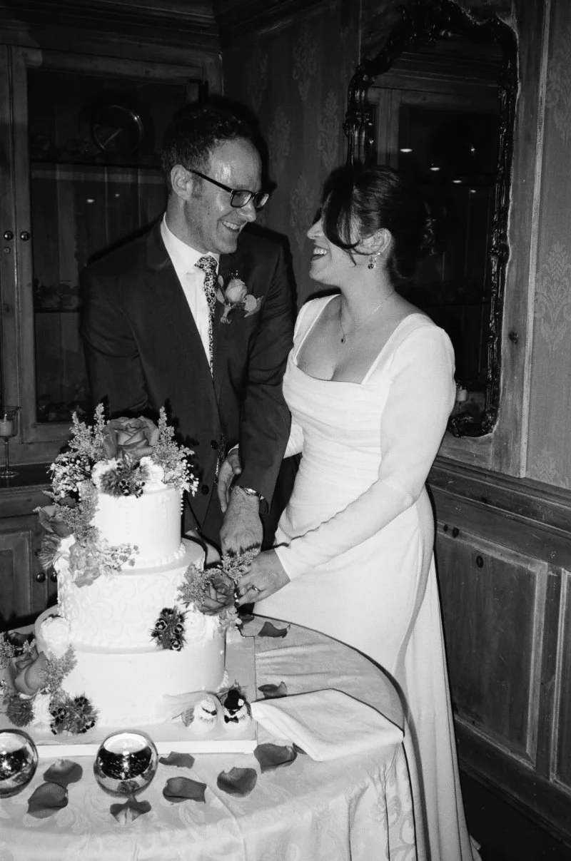 Bride and groom smiling while cutting floral wedding cake in black and white.