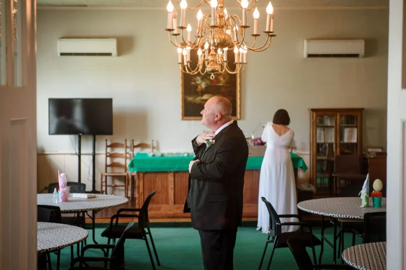 Wedding photography checklist moment featuring a groom getting ready while the bride prepares nearby.