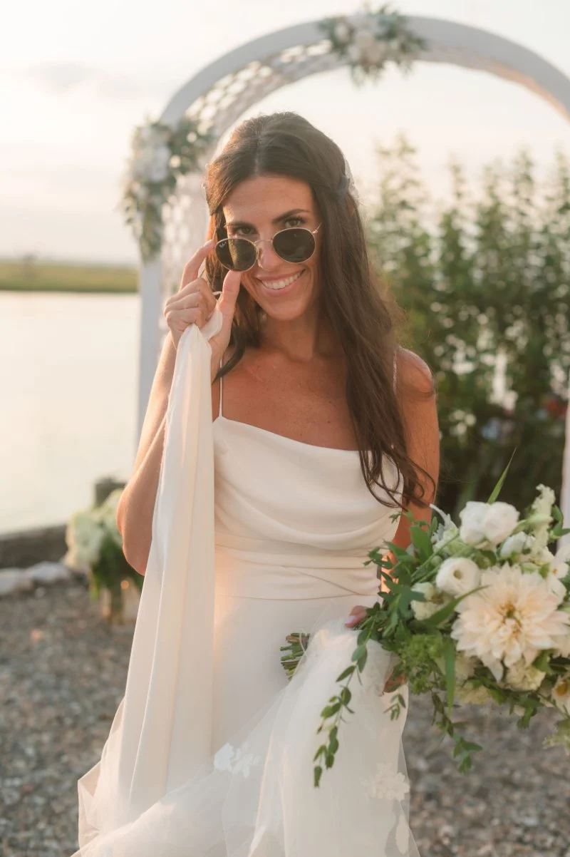 Bride smiling and adjusting sunglasses during golden hour photography beside a waterfront ceremony arch at sunset.