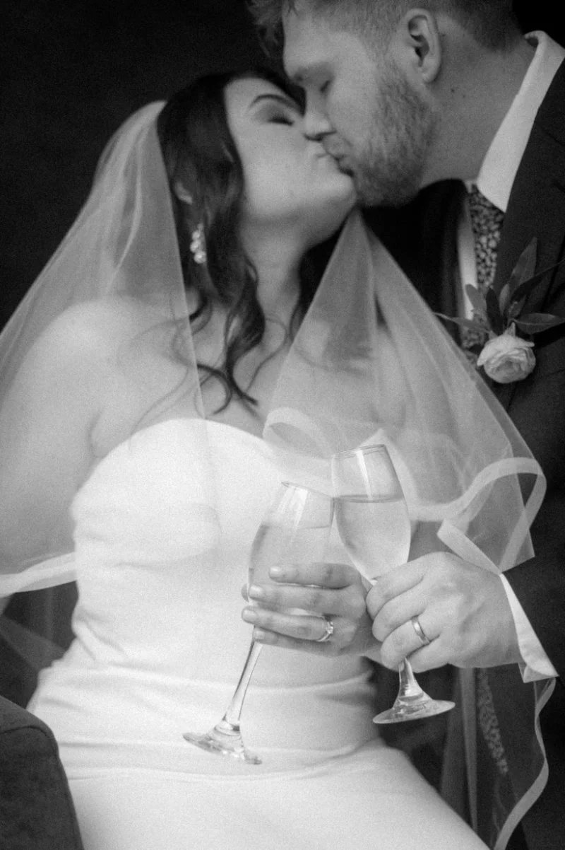 The couple kisses under a veil, holding champagne—quiet joy before the big wedding exit begins.