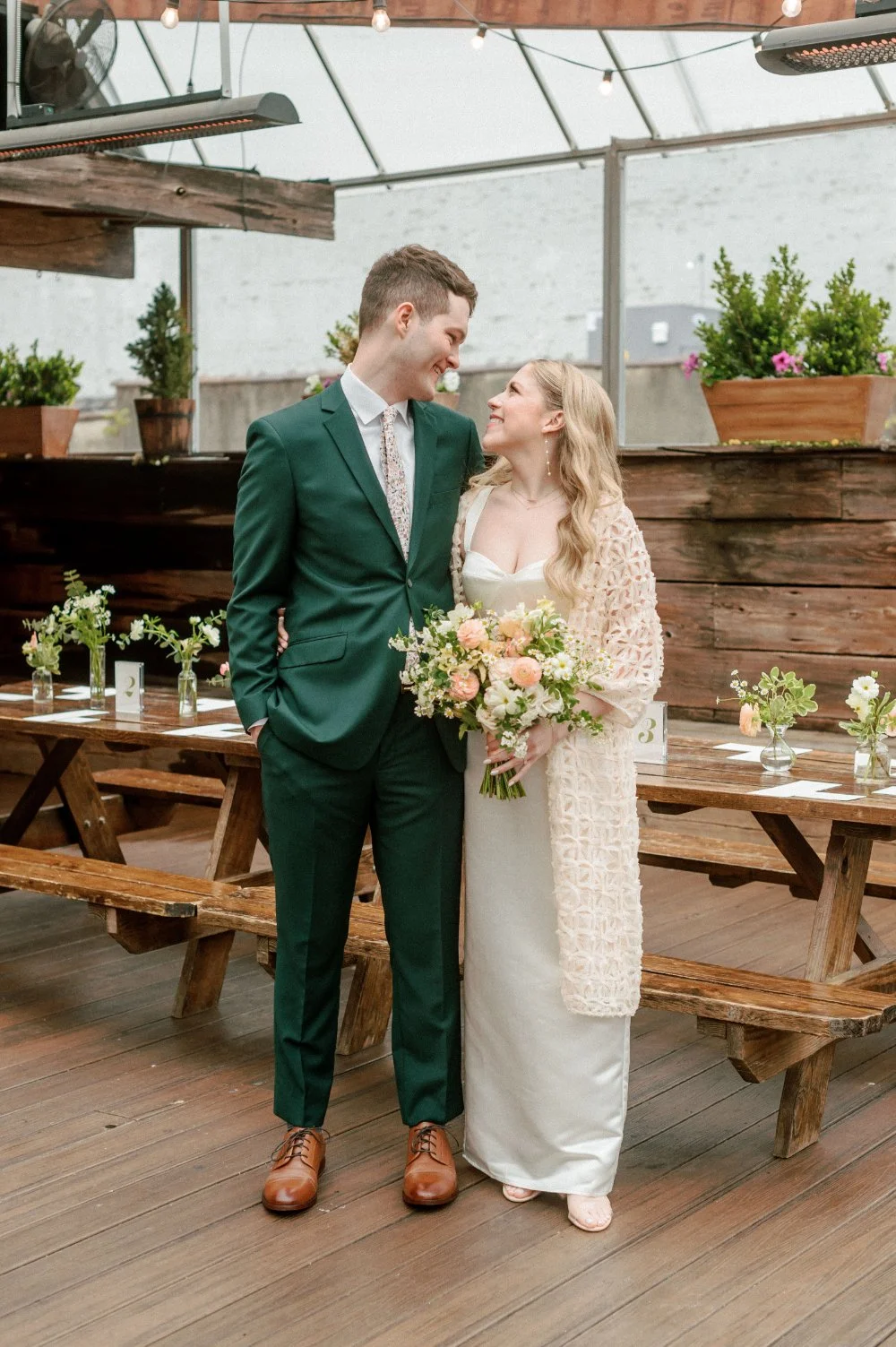 A joyful couple shares a tender look at their micro wedding in NJ & NYC, surrounded by rustic wood tables and soft florals, capturing warmth and intimacy in an airy venue setting.