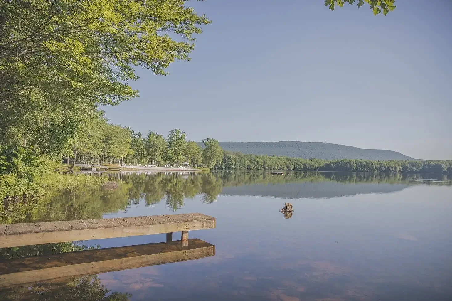 Calm lake with greenery along the shoreline, a small dock extending into the water, and distant hills under a clear blue sky.